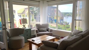 A living room with a sofa, armchair, and coffee table, large windows with blinds, and a view of houses and water. - Oceanside Retreat  Hubbards Nova Scotia (Hubbards)