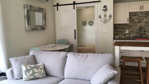 A modern living room with a grey sofa, a small table, and a kitchen area with white cabinets and a brick backsplash. - Oceanside Retreat  Hubbards Nova Scotia (Hubbards)