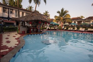 Une piscine avec des chaises longues et des parasols, un espace bar doté d’un toit de chaume, et des palmiers en arrière-plan.