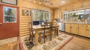 L’intérieur d’une cabane en rondins, avec une table à manger en bois, des chaises, une cuisine dotée de meubles en bois et une vue sur la forêt par les fenêtres.
