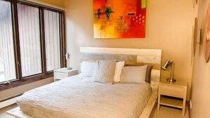 Een slaapkamer met een groot bed, een plafondventilator, een raam met zonwering en een kleurrijk abstract schilderij aan de muur.