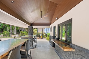 An outdoor patio with a stone wall, a wooden ceiling, and a ceiling fan. - Makena Surf • Bright and Airy Retreat • Steps to the Beach: Swim w/Turtles (Kihei)