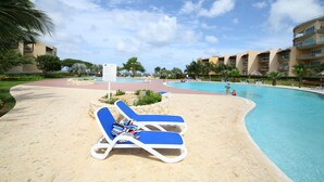 A poolside area with blue lounge chairs, a clear blue pool, and a building with balconies in the background. - 2 Bedroom, Beachfront, Amazing Ocean View, Modern ful (Eagle Beach)