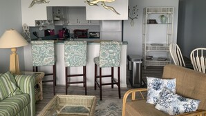 Eine moderne Küche mit weißen Schränken, einer Mikrowelle und einem Toaster. Drei Barhocker mit gemusterten Bezügen sind vorhanden. Im Wohnbereich befinden sich ein Korbstuhl und ein gestreiftes Sofa.