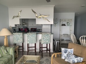 A modern kitchen with white cabinets, a microwave, and a toaster. There are three bar stools with patterned upholstery. A wicker chair and a striped sofa are in the living area.