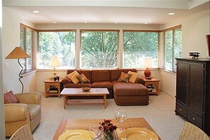 Relax in this inviting living room with a sofa, coffee table, and dining table for two, surrounded by large windows and a convenient cabinet.