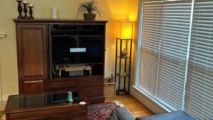 A living room with a dark wood entertainment center, a flat-screen TV, a glass coffee table, a gray sofa, and a window with blinds.