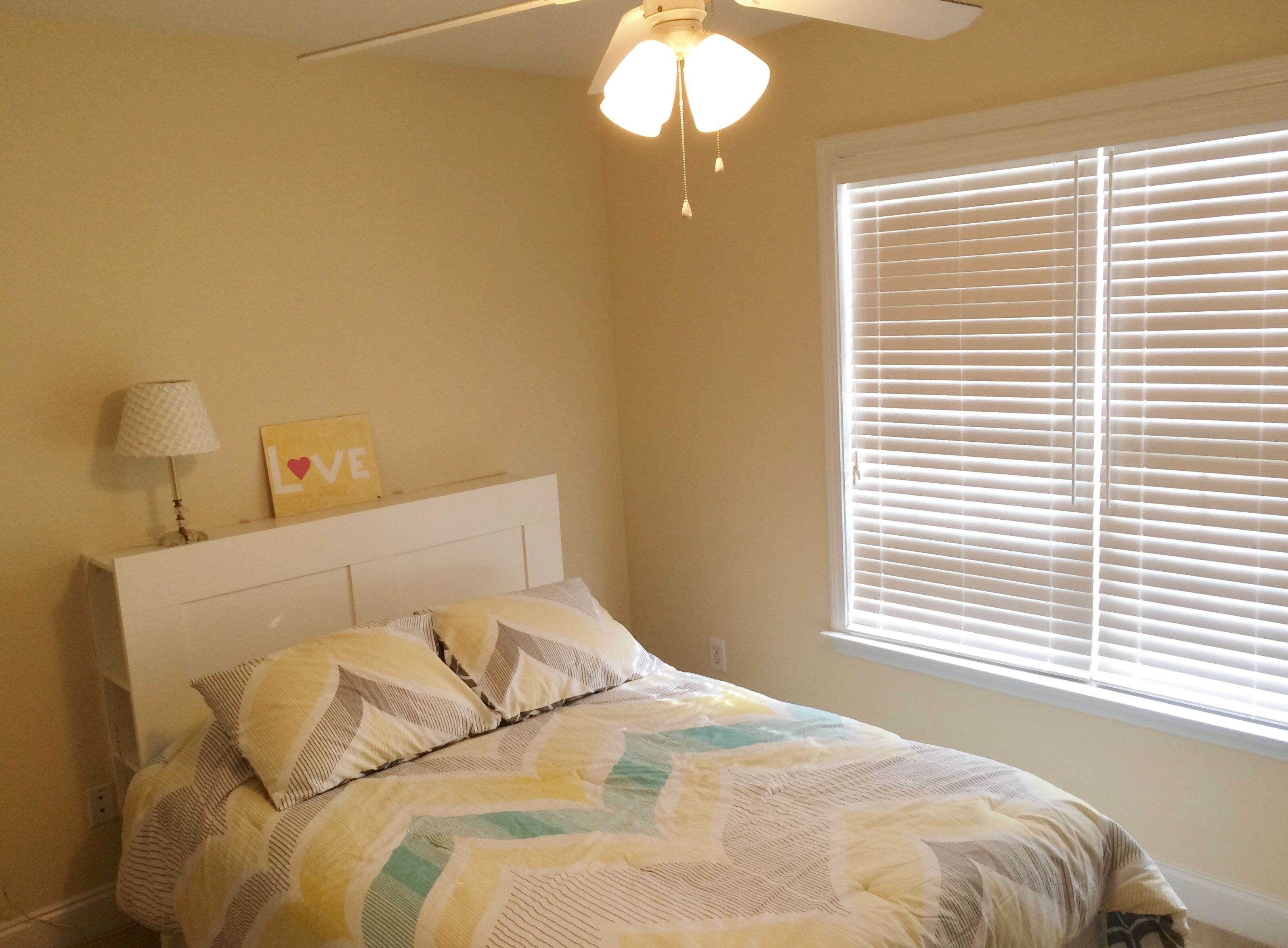 Une chambre à coucher avec un lit, un ventilateur de plafond, une fenêtre avec des stores et un mur orné d’un tableau encadré et d’une lampe.