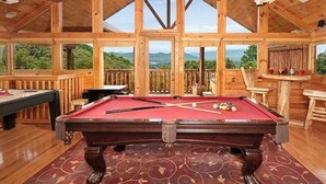 Una mesa de billar de madera en una habitación con ventanas grandes y vistas a las montañas.