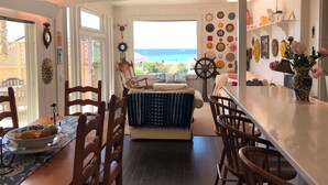 A cozy dining area with wooden chairs and a table, a view of the ocean through large windows, and various decorative items on the walls. - Last Chance! Unexpected opening– Steps to the Beach! Spectacular roof deck (Santa Cruz)