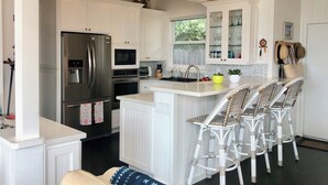 A modern kitchen with white cabinetry, a stainless steel refrigerator, a microwave, and a built-in oven. There is a bar area with wicker chairs and a small island with a sink. - Last Chance! Unexpected opening– Steps to the Beach! Spectacular roof deck (Santa Cruz)