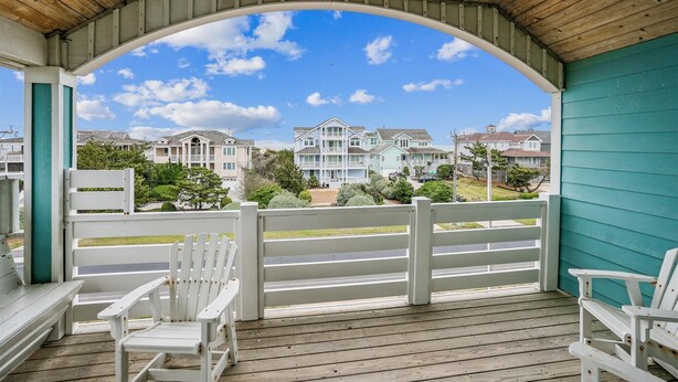 Un porche cubierto con sillas Adirondack blancas, vista a una zona residencial con casas y un cielo azul despejado.