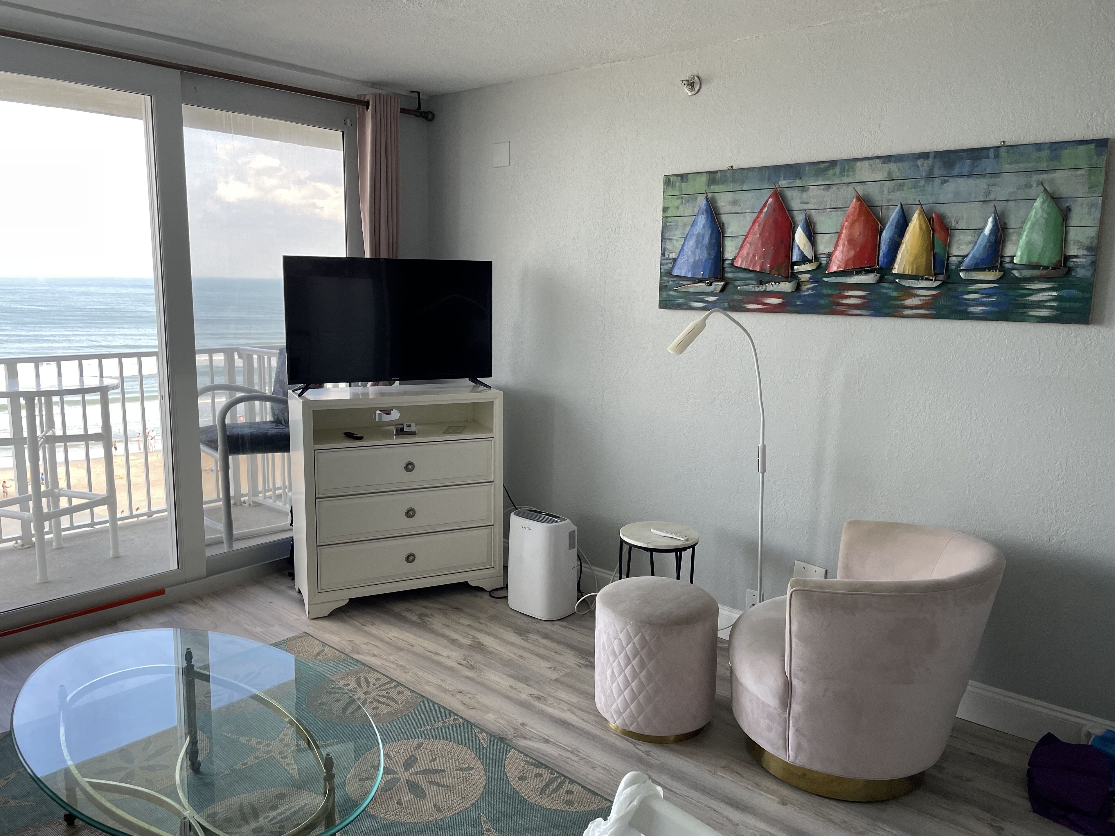 Une pièce avec une télévision posée sur une commode, un tableau représentant des voiliers, une table en verre et une vue sur la plage.