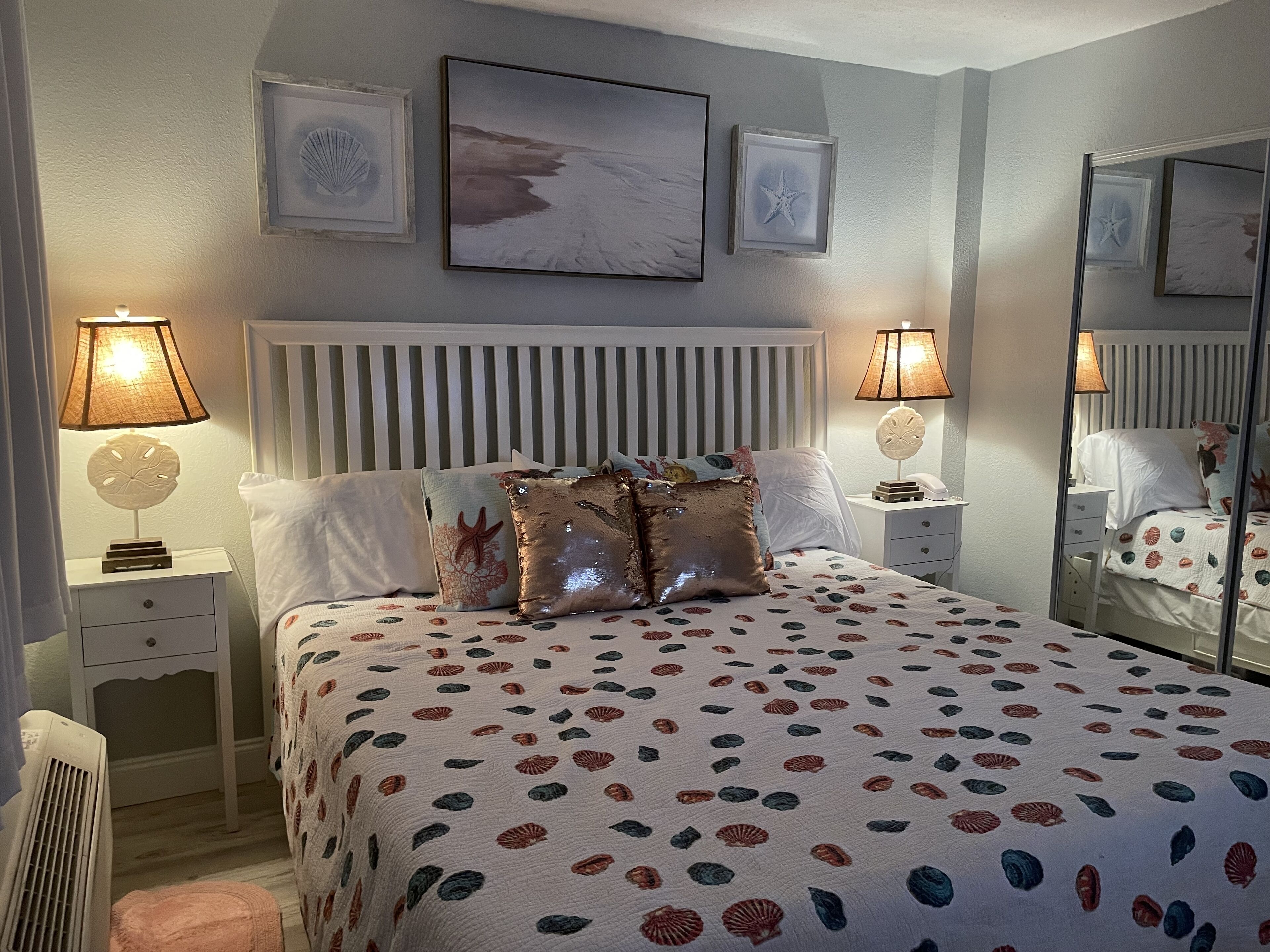 A bedroom with a bed, two bedside tables, a nightstand, a mirror, and wall art.