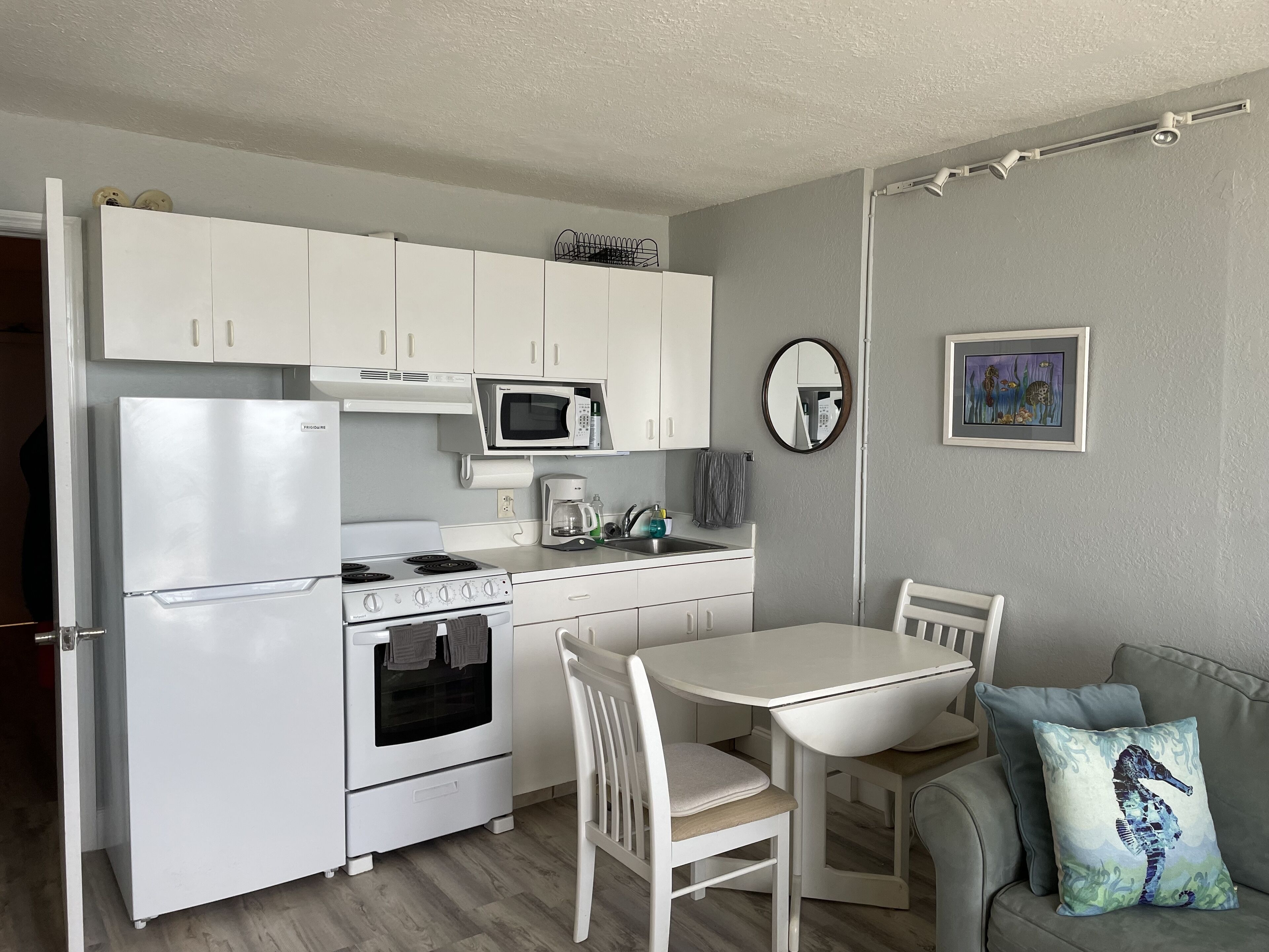 A compact kitchen with white appliances, a dining table, and a gray sofa with a cushion.