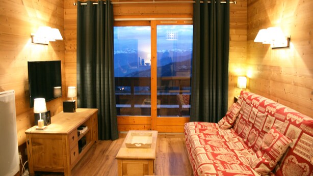 A cozy room with wooden walls, a wooden sofa with patterned cushions, a small wooden table, a TV on a stand, and a window with curtains. - Apartment ,Direct access to the slopes,4-6 persons,35 m2,beautiful view,wifi (Les Allues)