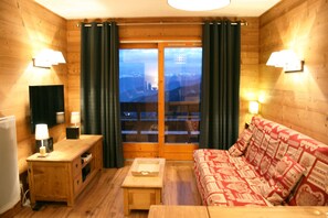 Una stanza accogliente con pareti in legno, un divano di legno con cuscini decorati, un tavolino e una televisione su un mobile, oltre a una finestra con tende.