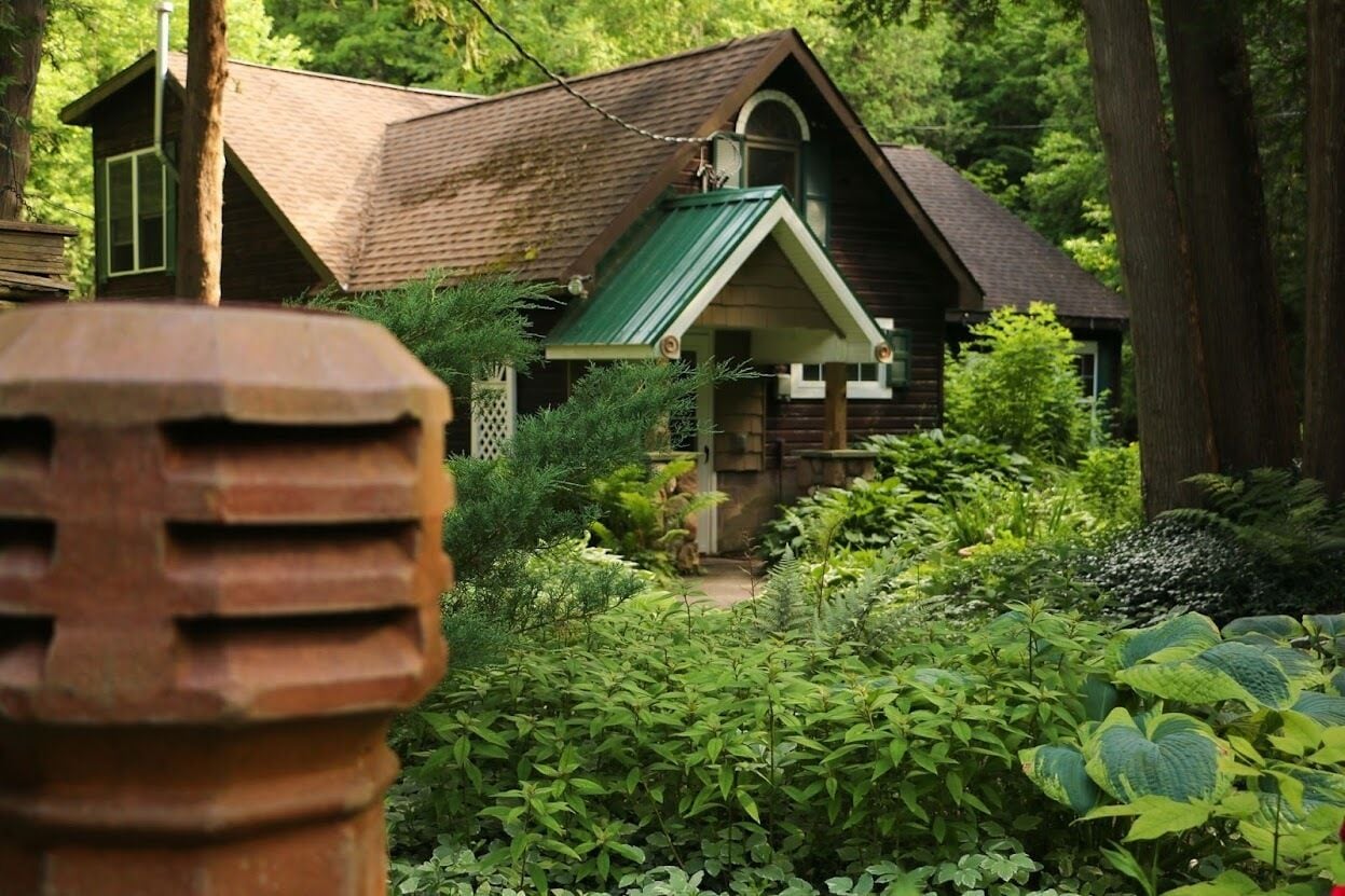 A rustic cabin with a green roof, surrounded by dense greenery and a wooden fence.