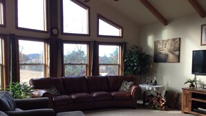 A cozy living room with a leather sofa, a patterned armchair, and a small table with decorative items. Large windows provide natural light and a view of the outdoors.