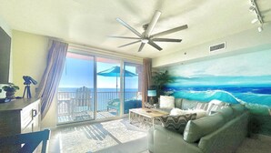 A living room with a balcony, a ceiling fan, a painting of the ocean, and a view of the sea. - Welcome to your Gulf Coast Paradise with Stunning Ocean Views & Extras Galore!!! (Panama City Beach)