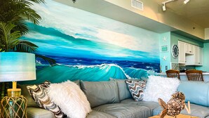 A living room with a large ocean mural, a grey sofa, a wooden coffee table, and a kitchen area in the background. - Welcome to your Gulf Coast Paradise with Stunning Ocean Views & Extras Galore!!! (Panama City Beach)