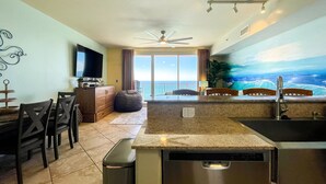 A modern kitchen with a built-in dishwasher, a dining area with chairs, a television, and a large window overlooking the ocean. - Welcome to your Gulf Coast Paradise with Stunning Ocean Views & Extras Galore!!! (Panama City Beach)
