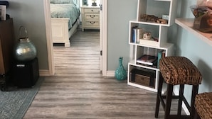 A room with a wooden floor, a bed with a green bedspread, a white dresser, a teal vase, and a wall clock.
