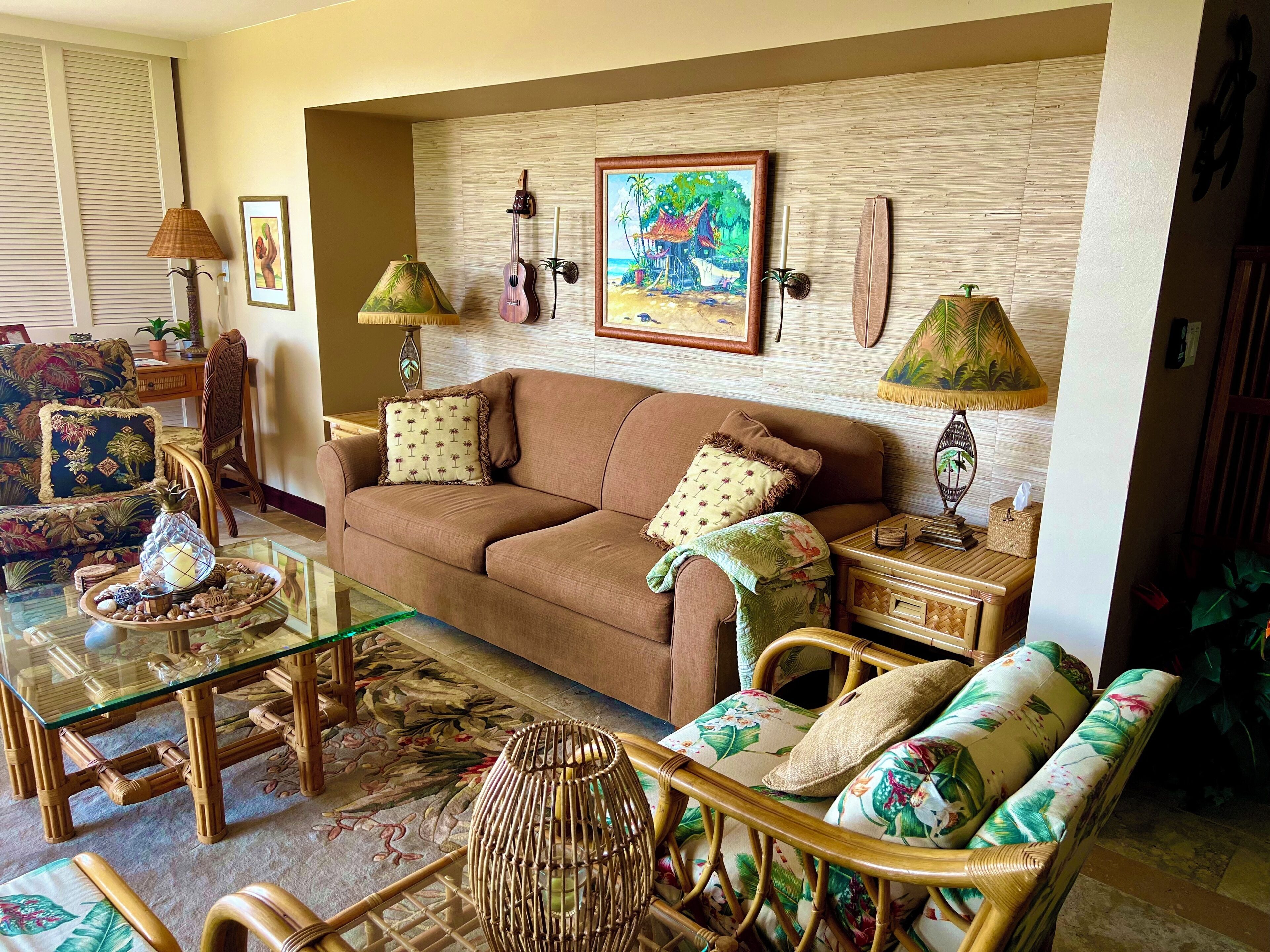 Un salon avec un canapé marron, une table basse en verre, des fauteuils en osier et un tapis à motifs.