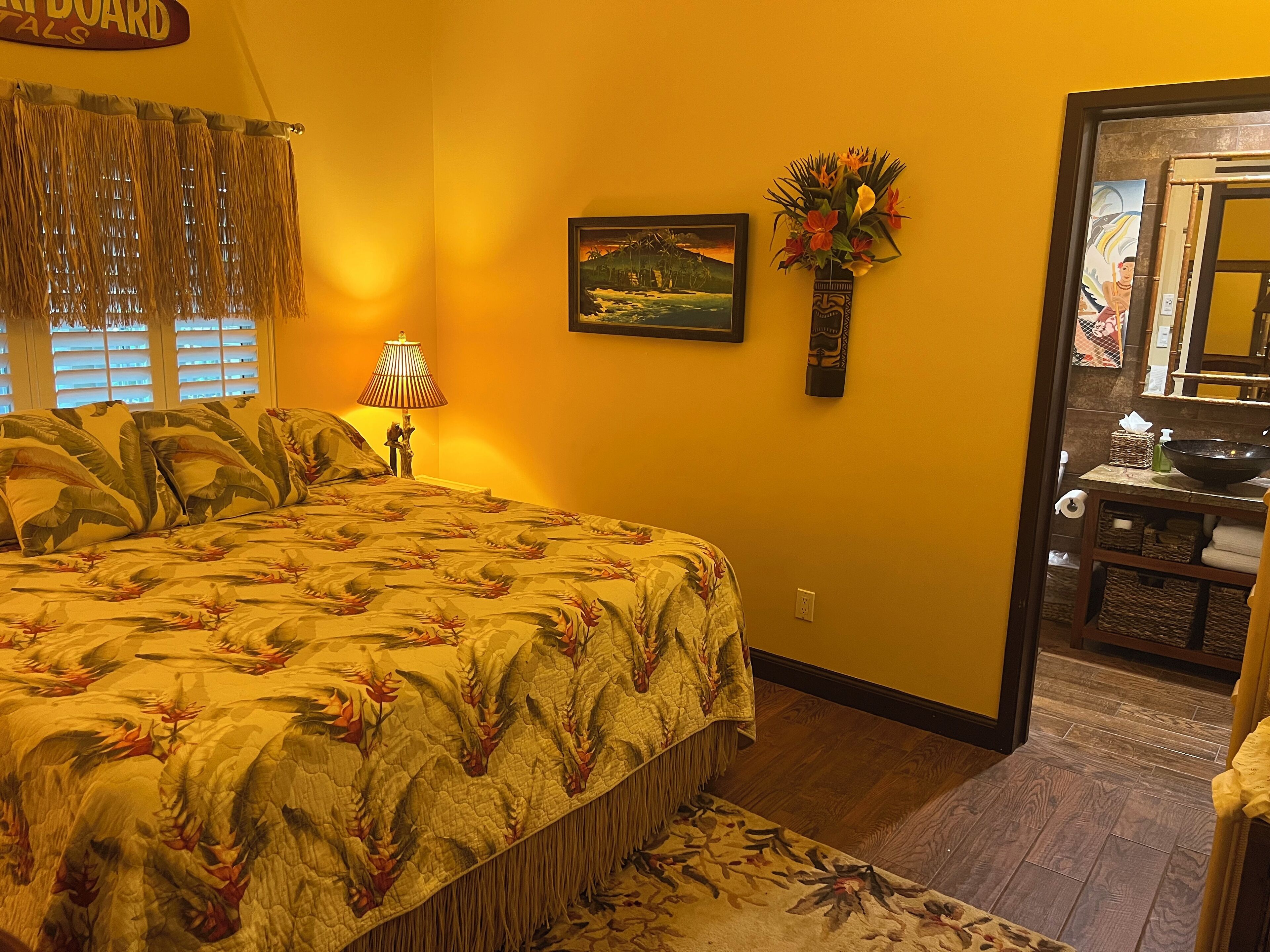 A room with a bed, a painting, a vase with flowers, and a bathroom visible through an open door.