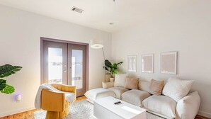 A modern living room with a sofa, a white coffee table, a plant, and a door leading to a balcony. - Walk to Stadium + GWCC | Modern 2BR w Parking (Atlanta)