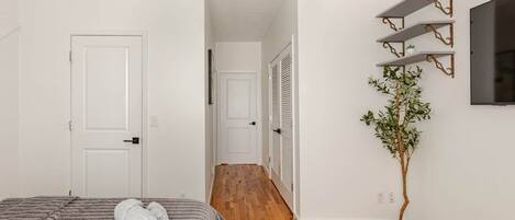 A bedroom with a bed, a bedside plant, a wall-mounted TV, and a hallway leading to another room.