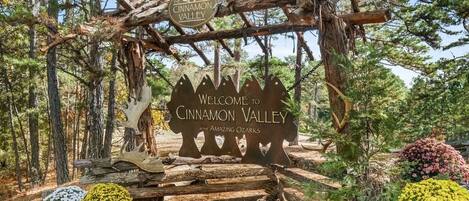 Un letrero de madera con la inscripción 'Welcome to Cinnamon Valley' y 'The Cinnamon Valley', rodeado de árboles y flores.