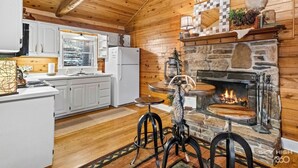 Interior rústico de una cabaña con chimenea de piedra, vigas de madera, cocina con armarios blancos y zona de comedor con taburetes.
