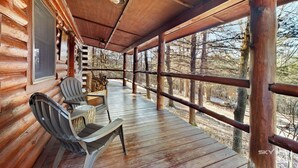 Una cabaña de madera con un porche cubierto, dos sillas Adirondack y un ventanal con vistas a un bosque.