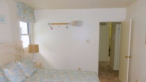 A bedroom with a bed, a nightstand, a lamp, a window with a curtain, a wall clock, and a door leading to another room.