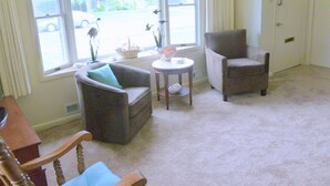 A living room with two armchairs, a small table, and a window with a view of a street and greenery.