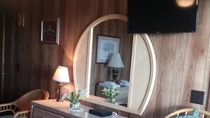 A room with wooden panel walls, a flat-screen TV mounted above a dresser, wicker chairs, a bed with a patterned bedspread, and a mirror with a round frame.