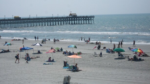 A beach with people, umbrellas, and a pier in the background.