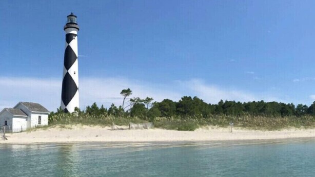A lighthouse with a distinctive black and white pattern, situated on a sandy beach with a clear view of the water.