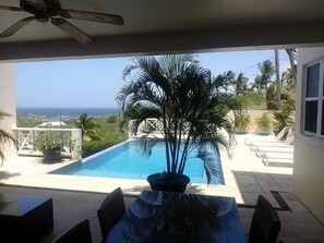 A pool area with a view of the ocean, a ceiling fan, and a table with chairs.