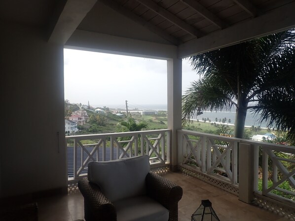 A balcony with a wicker chair, a view of a coastal area, and a palm tree.