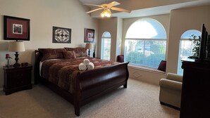 A bedroom with a large bed, a ceiling fan, and a view through the windows.