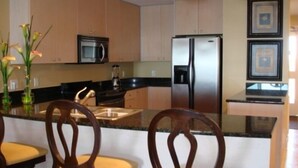 Eine moderne Küche mit Holzausstattung, einem Edelstahlkühlschrank, einer Mikrowelle und einer Arbeitsplatte aus Granit. Drei Holzstühle mit gepolsterten Sitzflächen sind vorhanden.