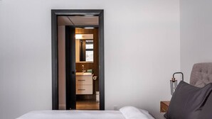 A bedroom with a bed, a nightstand, and a chair. There is a door leading to a bathroom with a mirror, sink, and toilet. - Tremblant Prestige - Horizon 1-104 (Mont-Tremblant)