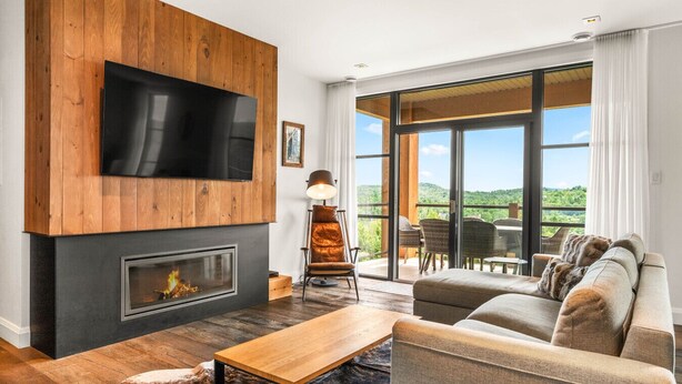 A modern living room with a fireplace, a flat-screen TV, and a large window offering a view of the outdoors. - Tremblant Prestige - Horizon 1-104 (Mont-Tremblant)