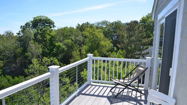 A wooden deck with a white railing, a single lounge chair, and a view of dense green trees in the background.