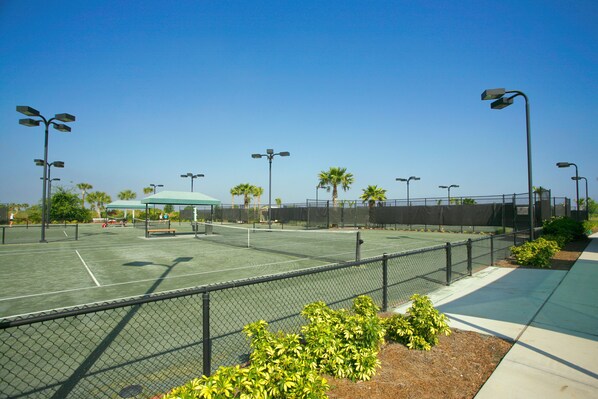 A tennis court with a chain-link fence, street lamps, and a covered seating area.