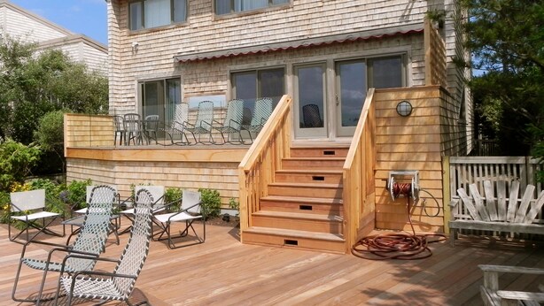 Ein zweistöckiges Holzhaus mit einer Terrasse, Sitzgelegenheiten im Freien und einer Treppe, die zu der oberen Etage führt.