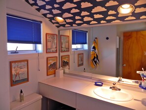 Ein Badezimmer mit einem weißen Waschbecken, einem Spiegel und einer Decke mit Wolkenmustern. An der Wand befinden sich Bilder in Rahmen und ein Handtuch hängt an der Tür.