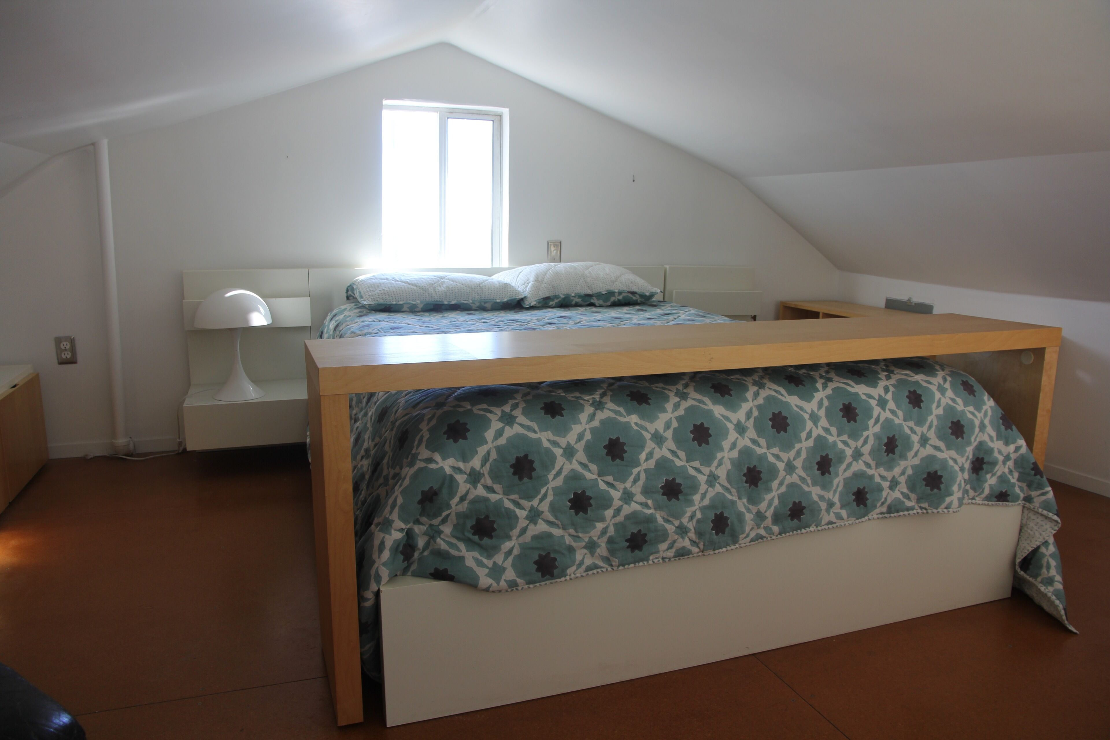 Una piccola camera con un letto singolo, un soffitto spiovente, una finestra e un comodino con una lampada.